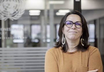 A woman in an orange shirt and black glasses stands in a United Nations office with her arms crossed. 