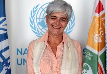 A woman smiles at the camera in front of the United Nations Sustainable Development Goals Flag and the United Nations Symbol.