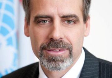 一个男人直视镜头，背景是联合国的旗帜。