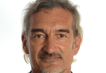 A man in a suit and tie looks directly at the camera with a white background. 