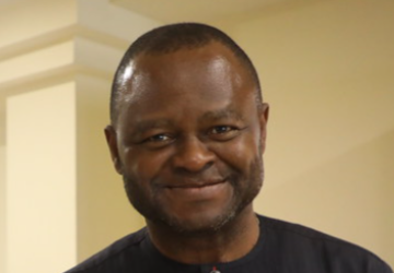 一名身穿黑色衬衫的男子对着镜头微笑，他站在一堵浅黄色的墙上。