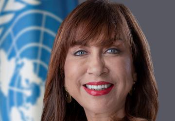 Women in white blazer smiles in front of UN flag 