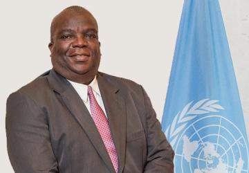 A man in a brown suit standing in front of the blue UN flag