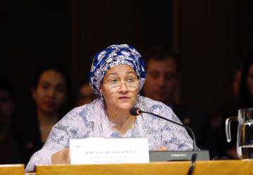 A woman in a lavender dress and deep purple headscarf speaks into a microphone