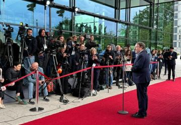 A man in a black suit stands at a podium on a red carpet and addresses a group of mediapersons with cameras and microphones