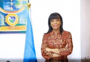 A woman with dark hair wearing a brown shirt with her arms crossed, stands in front of a white wall next to a blue UN flag
