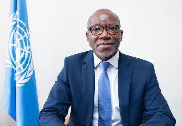 A man in a dark navy suit and blue tie with black spectacle frames sits in front of a blue flag with a white UN logo.