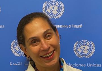 A woman in a white dress and colourful necklace leans against a blue background