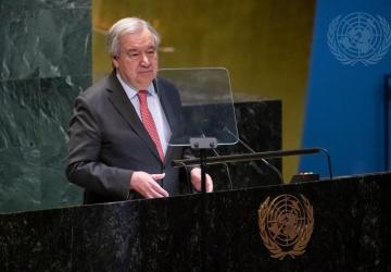 A man in a black suit and red tie stands at a podium and speaks into the microphone against a golden wall of the UN General Assembly in New York.