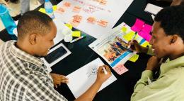 Deux jeunes hommes africains sont assis à un bureau de couleur noire couvert de post-its, de feuilles de papier et d'objets divers. Ils réfléchissent ensemble et esquissent des solutions sur une feuille de papier en souriant.