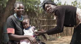 Une mère est assise avec sa petite fille tandis qu'une femme se penche sur fillette pour lui passer une petite bande autour du bras. Toutes trois regardent la caméra.