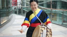 Une jeune fille vêtue d’un habit traditionnel se tient dans les allées d’un grand bâtiment moderne. Elle joue quelques percussions sur un instrument traditionnel, en soutiant.