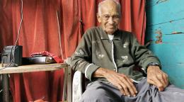 Candelario, un anciano, se sienta cerca de una mesa con equipos electrónicos. Él está mirando a la cámara, sonriendo.