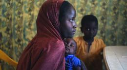 أم شابة تحمل طفلها النائم، بينما يظهر فتى يلبس قميصًا أصفر في الخلفية.