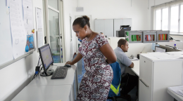 A woman in a dress works at a computer while a man near her works at his desk.