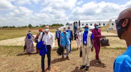 Varias personas con mascarillas, bolsas en la mano y chaquetas, descienden en un terreno en un día soleado. 