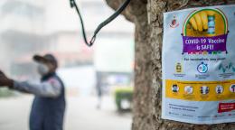 在一棵树上贴着一个标牌，上面有疫苗的图像和信息，可以看到背景中一个戴着面具的人在指点。