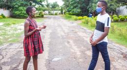 يقف فتى وفتاة يلبسان قناعي وجه متباعدان اجتماعيًا أثناء التحدث في منتصف الطريق في لوساكا، زامبيا.