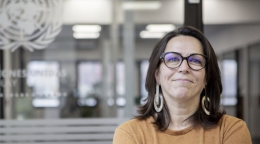 A woman in an orange shirt and black glasses stands in a United Nations office with her arms crossed. 