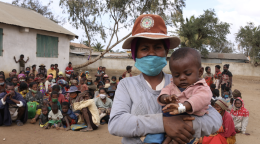A woman in a mask and a hat holds a baby in her arms while a large group of people sit behind her.