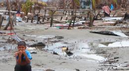 Un niño en primer plano, con un fondo de barro a sus espaldas, entre los escombros de lo que fue su hogar.