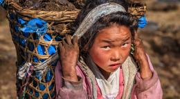 A young girl, with dirt on her hands and face, carries a large basket on her back.