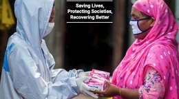 One healthcare worker wearing head-to-toe personal protective equipment hands a woman a handful of boxes. 