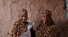Dos chicas sonríen sentadas en el suelo contra una pared mientras sostienen documentos.