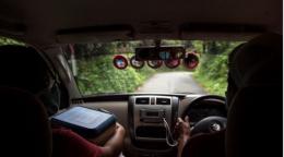 An illustrative photo from the backseat of a jeep in a green terrain, with one passenger carrying a box of what appears to be vaccines.