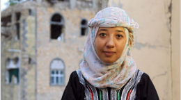 امرأة تجلس أمام مبنى مدمر في الخلفية.