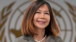 Una mujer con perlas y una camisa color canela se encuentra frente a un fondo con el logo de las Naciones Unidas.