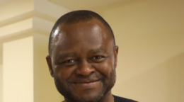 一名身穿黑色衬衫的男子对着镜头微笑，他站在一堵浅黄色的墙上。