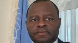 Un hombre se encuentra junto a una bandera de las Naciones Unidas y sonríe para la cámara. 