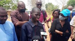 Un jeune adolescent béninois vêtu d'une blouse de mécanicien et entouré de plusieurs hommes et femmes répond aux micro de plusieurs journalistes en souriant. 