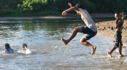 A group of young kids jump into a pool of water that appears like a lake with sand.
