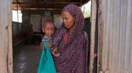 Woman in a brown dress holds a baby in blue as she stands in a doorway of a dilapidated room