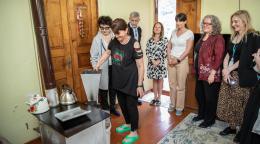 A group of people gather around a woman in a black shirt inside a house. The woman is putting shells into a silver heating machine.