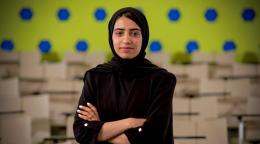 An Afghan girl in a black dress and headscarf stands in front of a polka dot wall