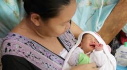 A woman in a purple dress cradles her new born baby in a white swaddle