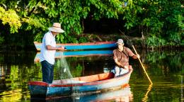Two fisherman on a small boat or canoe, on a body of water surrounded by greenery. 