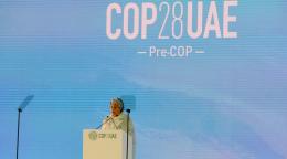 A UN official in a cream-colored outfit stands at a podium on a stage with a sign that says 'COP 28 UAE'.