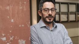 A man in a grey shirt and glasses stands next to a wall painted brown.