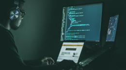 Мужчина в очках и наушниках, в темноте работает за компьютером и перед ним три разных экрана с информацией