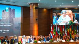 The deputy head of the UN sits on a panel with dignitaries and appears on a wide screen, with country's flags and flowers around the table.