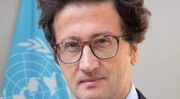 Mr. Joe Colombano wears a blue suite and red tie and stands in front of the UN flag