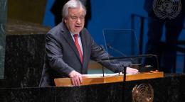 A man, the UN Chief, Antonio Guterres, in a black suit and red tie, stands at a podium against a blue background speaking into a microphone