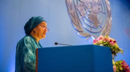 A woman in a green dress and headscarf speaks on a blue podium