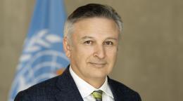 A man in a black suit and a green tie stands in front of a blue UN flaf