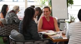 A woman in a red shirt talks to another woman in a black dress with other women in the background sitting around a table