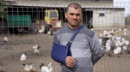 A man in a blue shirt and cast on his right arm stands in front of a grain barn and a chicken coop.
